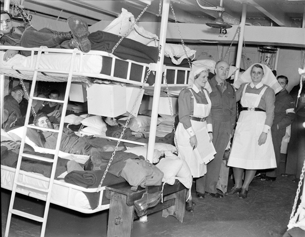Dans l'infirmerie du navire-hôpital, deux infirmières et un médecin debout devant des lits superposés où des soldats sont étendus.