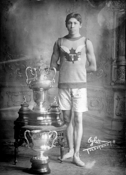 Jeune autochtone en costume de course, debout à côté de deux trophées.