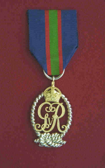Décoration pour Officiers de la Réserve Volontaire de la Marine Royale du Canada.  Une médaille ovale en argent doré chargée du chiffre royal GRI entouré d'une boucle de câble nouée en noeud plat au bas et sommée d'une couronne dorée.