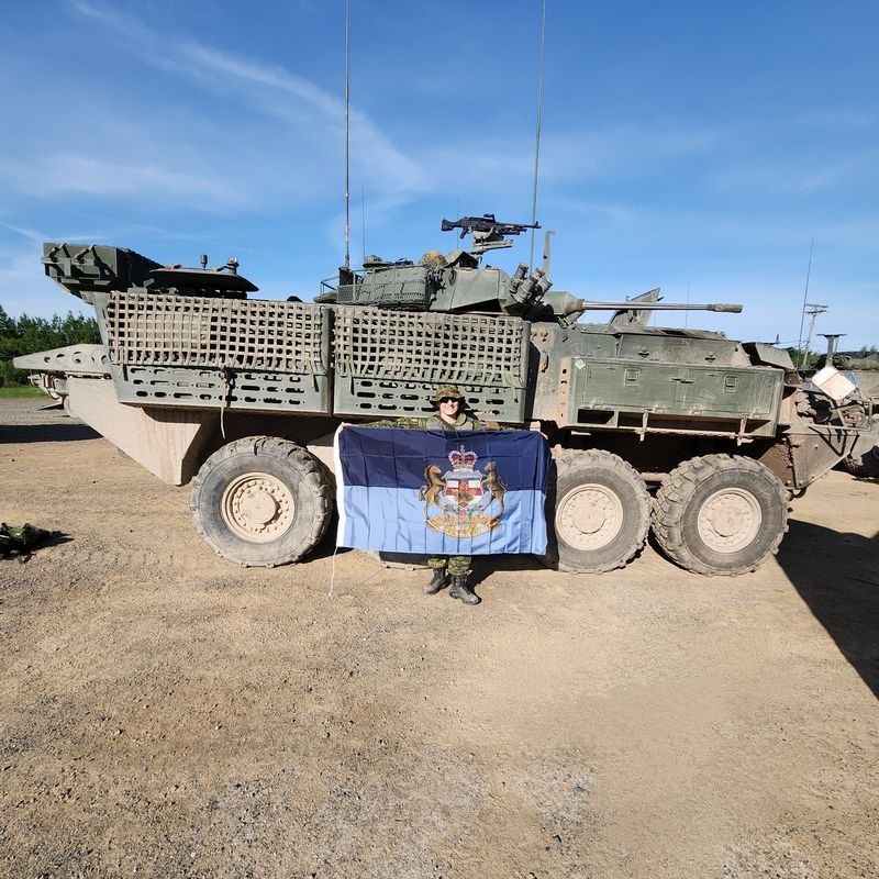 Shona Mitchell standing in front of a military vehicle. She is holding a blue flag with an emblem on it and is wearing a military uniform.
