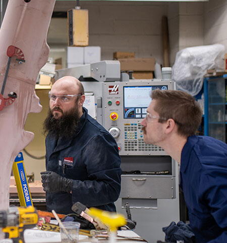 Deux personnes travaillent dans un atelier d’usinage à côté de gros modèles de troncs d’arbres roses, avec des outils et de l’équipement sur un établi.