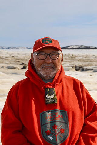 Solomon Awa standing in a field of snow and ice. He has a short beard and is wearing black-rimmed glasses and a red sweater with the Canadian Rangers logo on it.
